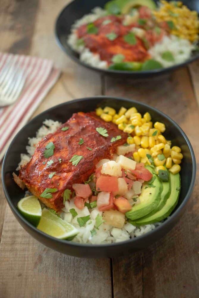 A filet of salmon on rice with avocado, salsa, corn and lime wedges