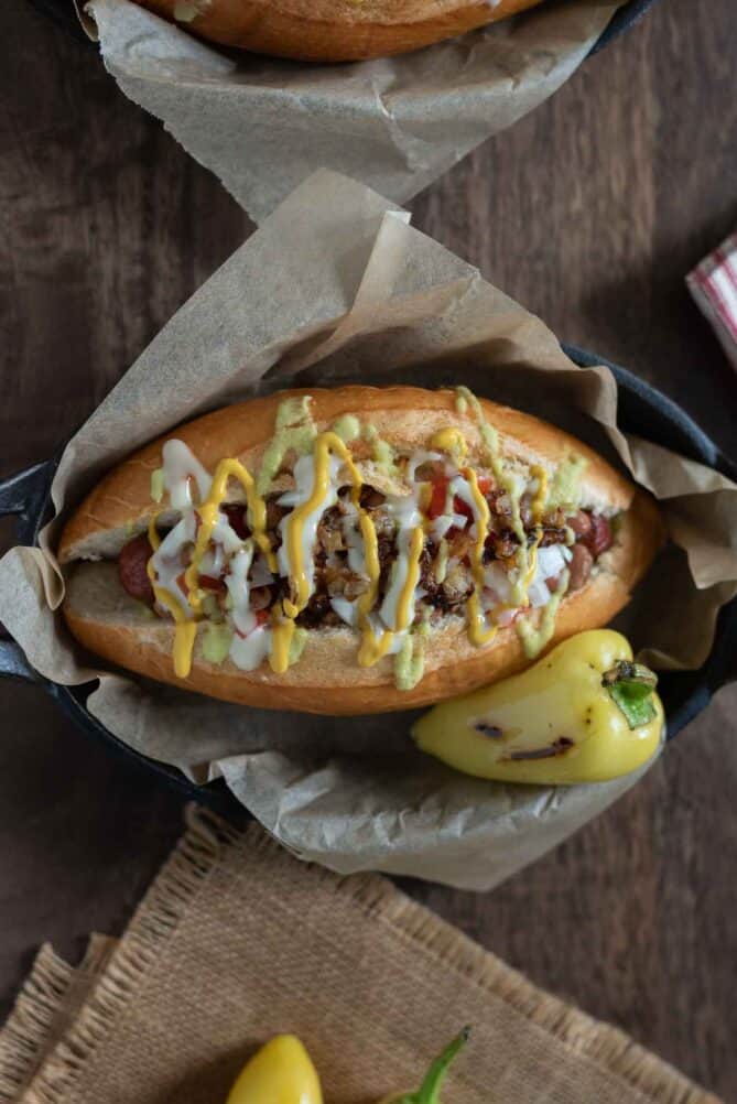 A bacon wrapped hot dog in a bun topped with pinto beans, onion and sauces