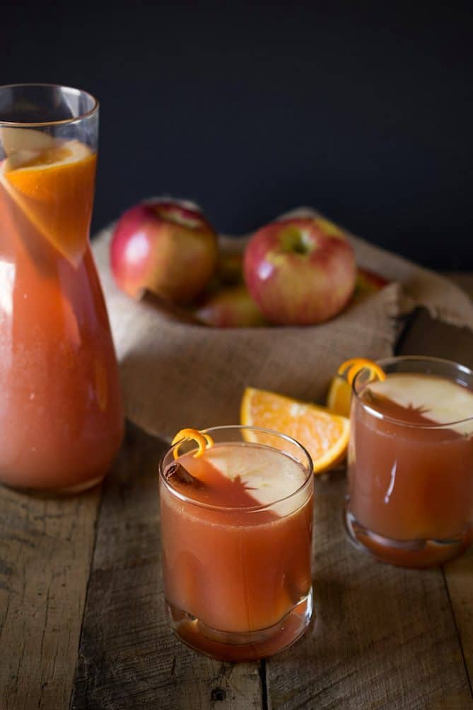 2 glasses of sparkling apple punch garnished with apple slice, cinnamon stock and orange zest with a pitcher and fresh apples in the background