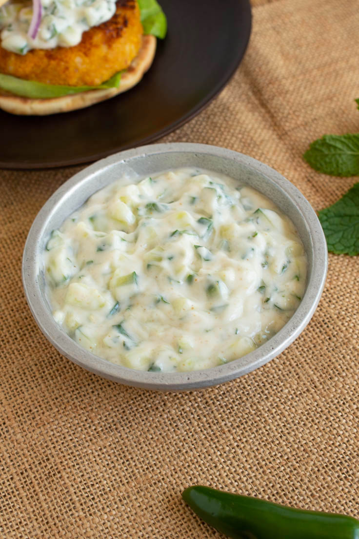 Cucumber raita in a silver bowl with a burger in the background