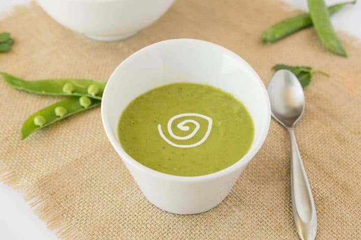 Vibrant green pea soup in a white garnished with sour cream with a spoon and fresh pea pods