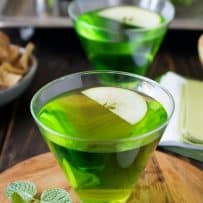 A very green drink in a stemless martini glass garnished with a fresh apple slice and minit