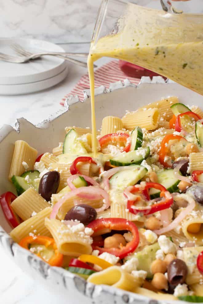 Creamy dressing being drizzled over a summer Greek pasta salad