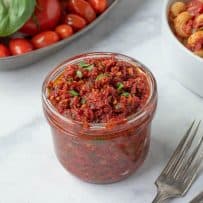 A jar of sun-dried tomato pesto with a bowl of pasta and fresh tomatoes and basil