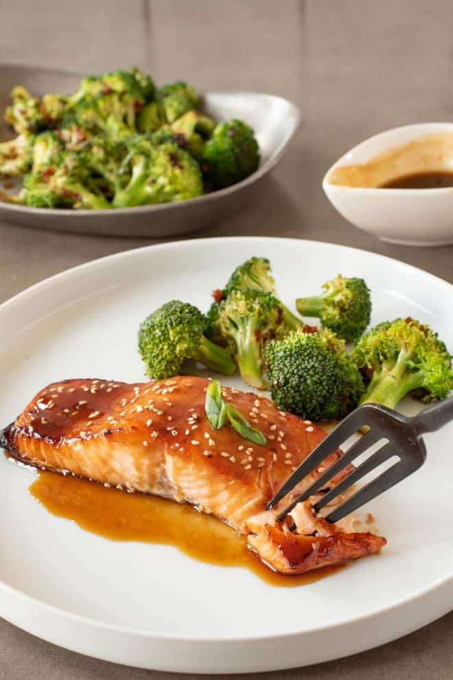 Using a fork to get a bite of teriyaki glazed salmon on a white plate with broccoli