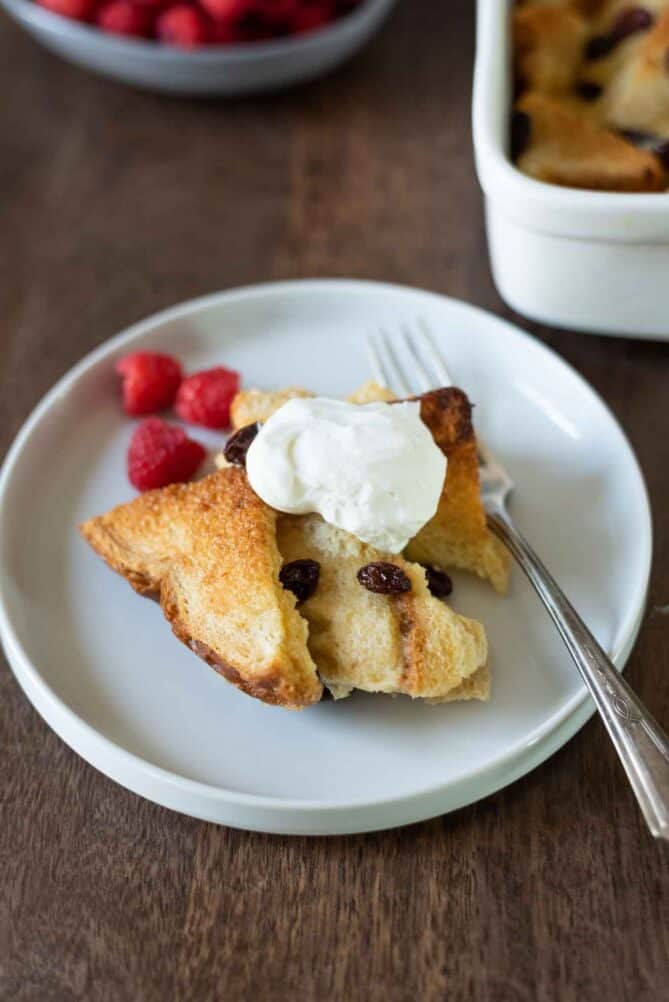 Bread and butter pudding served on a white plate topped with whipped cream and raspberries