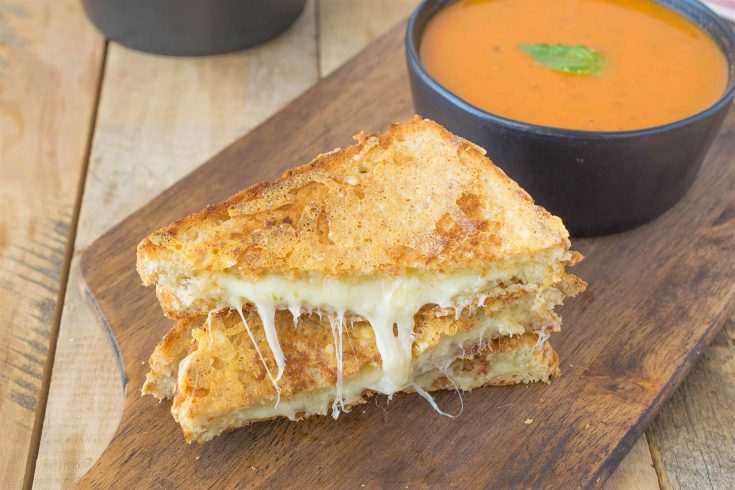 A&nbsp;triple grilled cheese sliced in half showing the melted cheese inside on a serving board with tomato basil soup
