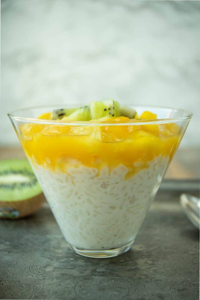 A glass filled with rice pudding viewed from the side showing the rice in the creamy sauce topped with sauced mango and pineapple
