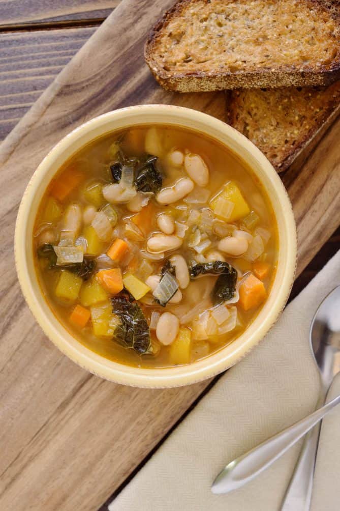 Tuscan bean soup viewed from overhead in a bowl with 2 spoons and 2 pieces of bread
