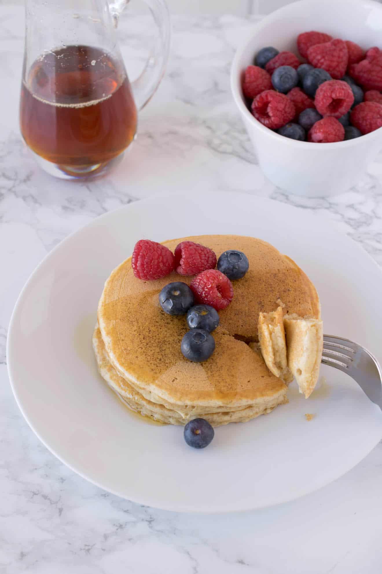 A plate of whole wheat pancakes with a bite taken out with fruit and syrup