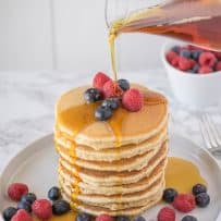 Pouring syrup of a large stack of pancakes surrounded with raspberries and blueberries